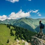 Adventurous hiker enjoying breathtaking views of lush green mountains in Romania under a clear blue sky.