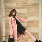 Fashion model in pink coat and black dress striking a dynamic pose indoors with stylish black heels.