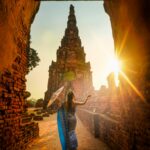 A woman in traditional wear stands with an umbrella at a historic Ayutthaya temple during sunset.