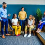 A diverse group of five professionals engaging in a casual indoor meeting, smiling and talking.