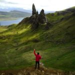 A lone hiker in a red jacket stands before the iconic Old Man of Storr on the Isle of Skye, Scotland.
